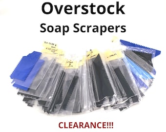 May include: A collection of soap scrapers in individual clear plastic sleeves, featuring various black and blue plastic tools with serrated edges. Each sleeve has a small adhesive label with handwritten text identifying the specific scraper design.
