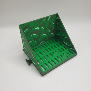 May include: A dark olive green angled shelf or rack featuring a bright green slotted base and a back panel decorated with a repeating light green and white leaf pattern. The structure has a solid frame with a small protruding tab on the left side.