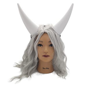 May include: A mannequin head with gray wavy hair and large white horns. The horns are curved and rise upwards. The mannequin has a neutral expression and the text "Hair Way" is printed on the neck.