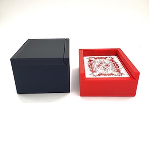 May include: A black rectangular box with a hinged lid sits next to a red rectangular card holder. The card holder contains a deck of playing cards with a red and white design.