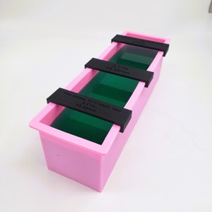 May include: A pink rectangular soap mold with three black crossbars. The mold contains green liquid. The text on the crossbars reads "Fits across Mold width size 3.41in 86.65mm".