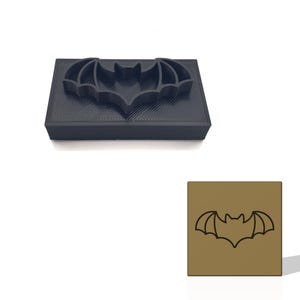 May include: A black bat-shaped cookie cutter with a rectangular base. The bat design is raised and has detailed wing and head features. A second image shows a bat outline on a brown square.
