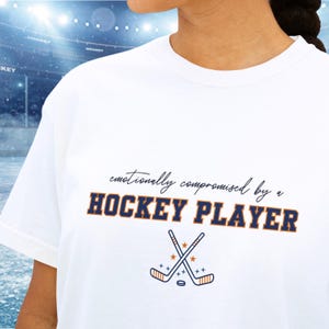 Puede incluir: Una camiseta blanca con el texto en cursiva negra "emotionally compromised by a" sobre el texto en negrita y letras de bloque azul oscuro "HOCKEY PLAYER" con un borde naranja. Debajo del texto hay dos palos de hockey cruzados y un disco.