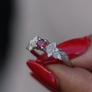 Op de afbeelding: Een zilveren ring met een rode edelsteen en drie gegraveerde rozenontwerpen.