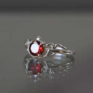 May include: A silver ring with a red gemstone and a smaller clear gemstone. The ring has a delicate, intricate design.