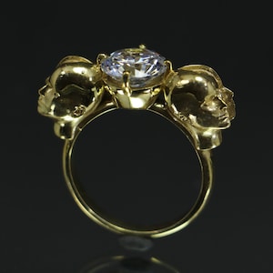 Puede incluir: Anillo de oro con tres caras estilizadas y una gran piedra preciosa transparente en el centro.