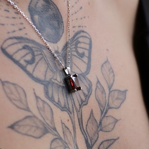 Puede incluir: Un collar de plata con un colgante rectangular de piedra preciosa rojo oscuro. El colgante está engastado en un bisel de plata y cuelga de una delicada cadena. El collar se muestra en el pecho de una persona, con un tatuaje visible en el fondo.