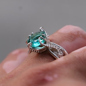 Pode incluir: Um anel de prata com uma pedra preciosa ovalada de cor turquesa. O design do anel incorpora tentáculos de polvo que envolvem a pedra. Os tentáculos estendem-se ao longo da banda, criando uma joia única e detalhada.