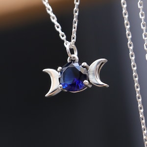 Puede incluir: Un collar de plata con un colgante de triple luna con una piedra preciosa azul. La cadena es de estilo delicado y de eslabones.