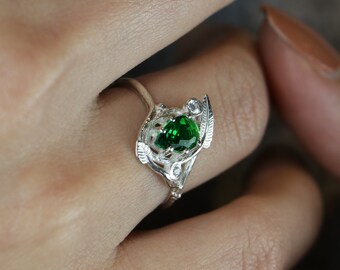 May include: A silver ring featuring a large, round, emerald-green gemstone as the focal point. The ring has an ornate, nature-inspired design with delicate leaf and feather details. A smaller, clear gemstone accents the design.