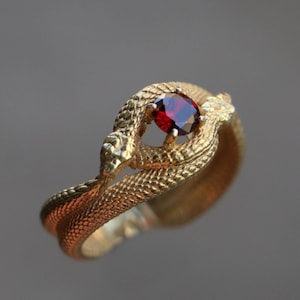 Puede incluir: Un anillo de oro con una piedra preciosa roja en el centro, con forma de dos serpientes entrelazadas.