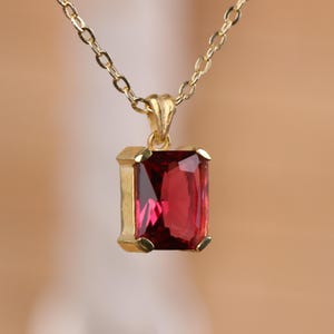 Op de afbeelding: Een goudkleurige ketting met een rechthoekige, dieprode edelsteen hanger. De hanger is gezet in een gouden frame en hangt aan een fijne gouden ketting. De edelsteen heeft een facetgeslepen oppervlak, dat licht reflecteert.