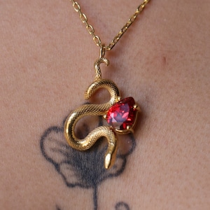 Peut inclure: Un collier de serpent en or avec un pendentif en pierre rouge. Le serpent est enroulé et la pierre est en forme de cœur.