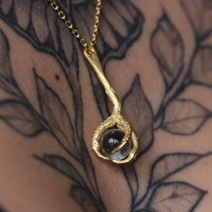 May include: A gold necklace with a claw pendant holding a clear glass orb. The necklace is on a person's chest with a tattoo of leaves and flowers.
