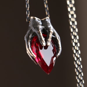 Könnte beinhalten: Eine silberne Halskette mit einem Anhänger, der zwei Hände zeigt, die einen großen, roten herzförmigen Edelstein halten.