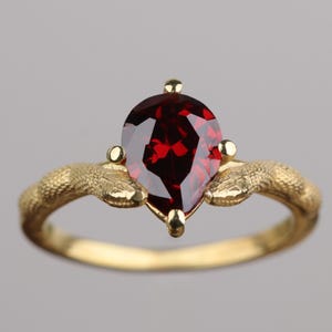 May include: A gold ring featuring a pear-shaped, deep red gemstone. The ring's band has a textured, organic design, with two snake-like elements flanking the gemstone. The ring is set against a neutral gray background.