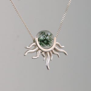K&ouml;nnte beinhalten: Eine silberne Halskette mit einem sonnenf&ouml;rmigen Anh&auml;nger. Der Anh&auml;nger hat eine klare, runde Mitte mit gr&uuml;nen Einschl&uuml;ssen, eingebettet in ein silbernes Sonnendesign. Die Halskette ist ebenfalls aus Silber.