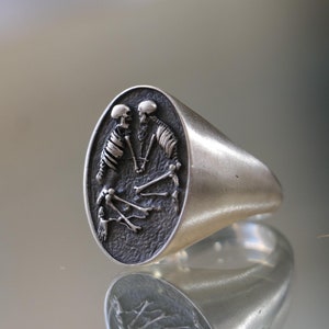 May include: A silver ring with a detailed design of two skeletons embracing. The ring is oval shaped and has a smooth, polished finish.
