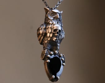 May include: A silver-toned owl pendant necklace. The owl has detailed feathering and a skull design on its chest. A large, black, teardrop-shaped stone is at the bottom, surrounded by small silver beads. The necklace chain is also silver.