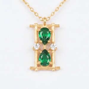 Puede incluir: Collar dorado con colgante. El colgante tiene dos gemas verdes en forma de pera y dos pequeñas gemas blancas. El colgante tiene un marco rectangular. El collar se muestra sobre un fondo blanco.