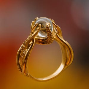 Puede incluir: Un anillo dorado con una gema esférica y transparente. El diseño del anillo presenta detalles intrincados y texturizados que sostienen la gema. La banda del anillo tiene un acabado liso y pulido, creando un contraste con el engaste texturizado.