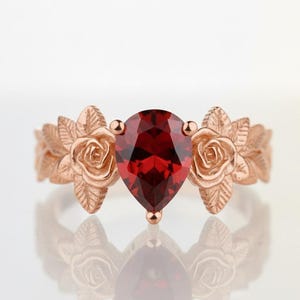 Könnte beinhalten: Roségoldener Ring mit einem tropfenförmigen, tiefroten Edelstein. Der Ring ist mit Rosen- und Blattmotiven verziert, die ein florales Design bilden. Der Edelstein ist der Blickfang, reflektiert das Licht und verleiht Eleganz.
