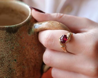 May include: A gold ring featuring a prominent oval-cut deep red gemstone set in a textured metal band. The ring is worn on a finger, showcasing the faceted ruby-colored stone and the intricate, organic-looking metalwork of the band's setting.