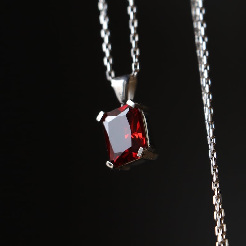 Pu&ograve; includere: Una collana con pendente in argento con una gemma rettangolare rosso scuro. La gemma &egrave; incastonata in una lunetta d'argento e pende da una delicata catena d'argento. Lo sfondo &egrave; nero.