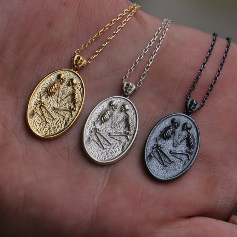 May include: Three silver oval pendants with a skeleton couple design. The pendants are on chains and are in different finishes: gold, silver, and black.