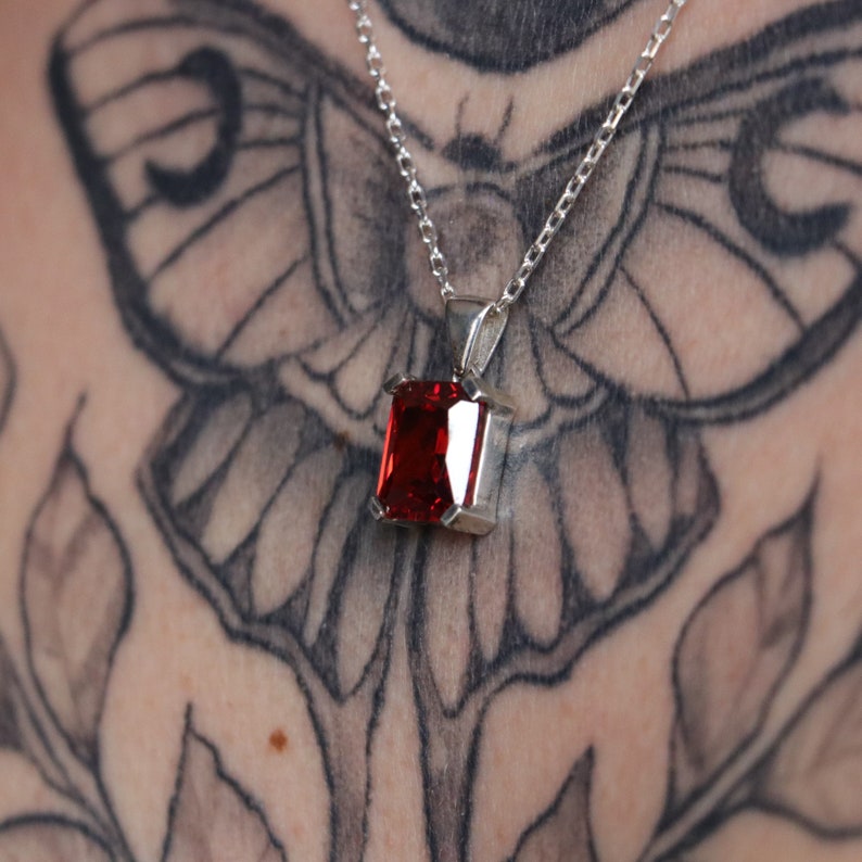 Pu&ograve; includere: Una collana d'argento con un pendente rettangolare di pietra preziosa rosso intenso. Il pendente &egrave; incastonato in una lunetta d'argento e pende da una delicata catena d'argento. La collana &egrave; mostrata su una pelle tatuata.