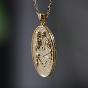 May include: A gold pendant necklace with an oval-shaped charm featuring a detailed design of two skeletons holding hands. The charm has a textured surface and a gold border.