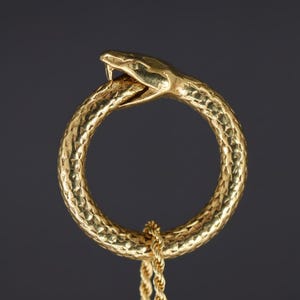 Op de afbeelding: Een goudkleurige ring in de vorm van een slang die in zijn eigen staart bijt, met een getextureerd oppervlak. Een gouden ketting is aan de onderkant van de ring bevestigd. De kop van de slang is gedetailleerd met open kaken en een zichtbare tong.