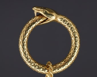 May include: A gold-colored ring in the shape of a snake biting its own tail, with a textured surface. A gold chain is attached to the bottom of the ring. The snake's head is detailed with open jaws and a visible tongue.