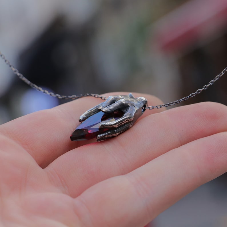 May include: A silver necklace with a hand holding a red gemstone pendant.