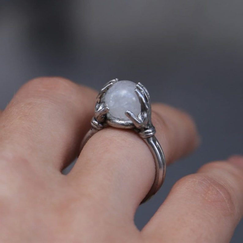 May include: A silver ring featuring a white moonstone held by two hands. The ring is worn on a finger.