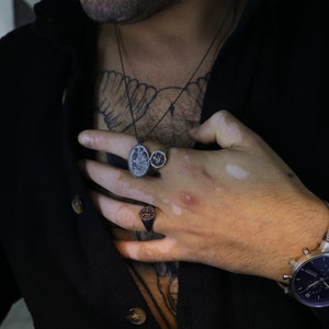 May include: A silver chain necklace with a black oval pendant featuring a detailed design of two birds. The necklace is worn by a person with a tattoo on their chest. The person is also wearing two silver rings on their finger.