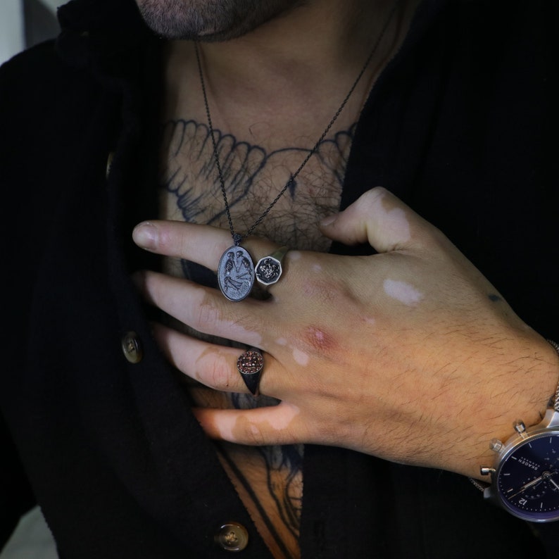 May include: A silver chain necklace with a black oval pendant featuring a detailed design of two birds. The necklace is worn by a person with a tattoo on their chest. The person is also wearing two silver rings on their finger.