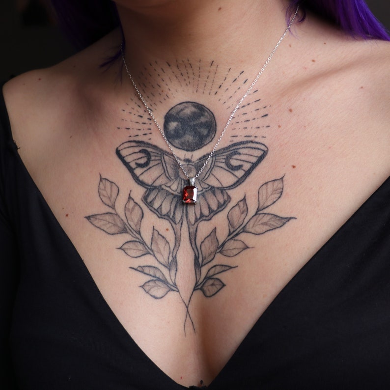 Pu&ograve; includere: Una collana d'argento con un pendente rettangolare di pietra rossa. Il pendente &egrave; appeso a una sottile catena d'argento. La collana &egrave; presentata su un top nero e un tatuaggio sul petto raffigurante una falena, foglie e una luna.