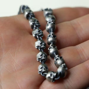 May include: A silver chain necklace with multiple skull-shaped beads. The necklace is made of metal and has a dark, aged finish.
