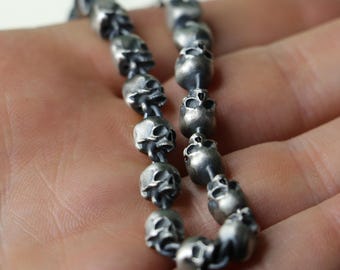 May include: A silver chain necklace with multiple skull-shaped beads. The necklace is made of metal and has a dark, aged finish.