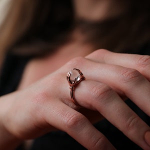 May include: A rose gold ring featuring a clear stone held by two hands. The ring is worn on a person's finger.