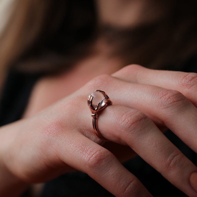 May include: A rose gold ring featuring a clear stone held by two hands. The ring is worn on a person's finger.