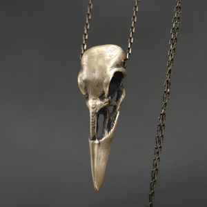 May include: A gold-toned pendant necklace featuring a detailed bird skull charm. The chain is made of a dark metal.