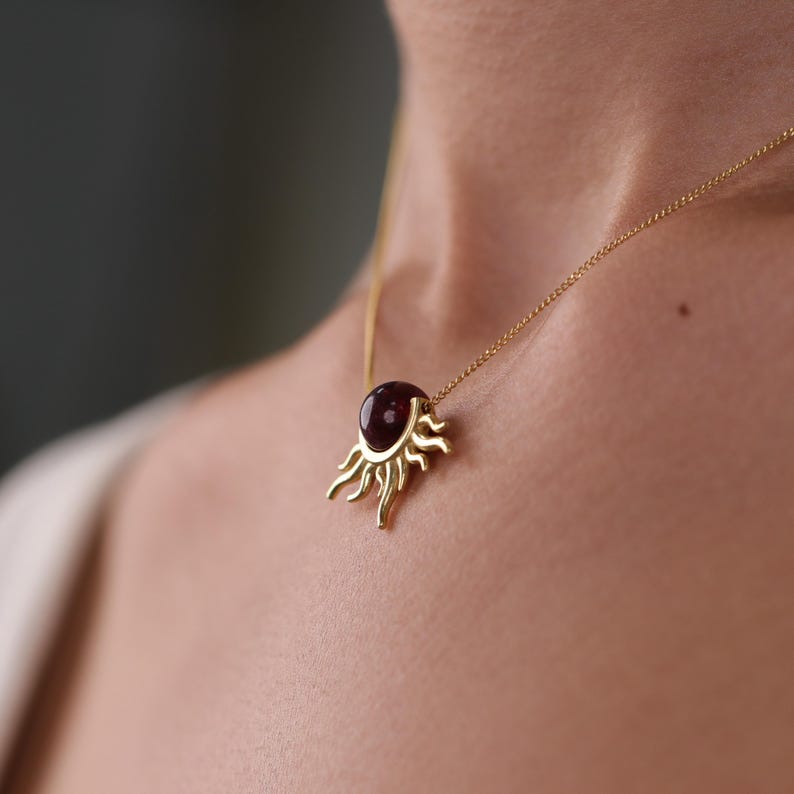 K&ouml;nnte beinhalten: Eine goldfarbene Halskette mit einem sonnenf&ouml;rmigen Anh&auml;nger. Der Anh&auml;nger hat einen gro&szlig;en, ovalen, dunkelroten Edelstein in der Mitte, umgeben von stilisierten Strahlen. Die Halskette wird auf der Haut einer Person getragen.