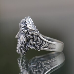 May include: Silver ring with a detailed sculpted design of a man's face wearing a laurel wreath. The ring is shown from the side.