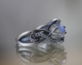 May include: A silver ring with an intricate design featuring an octopus tentacle wrapping around a large, iridescent moonstone.