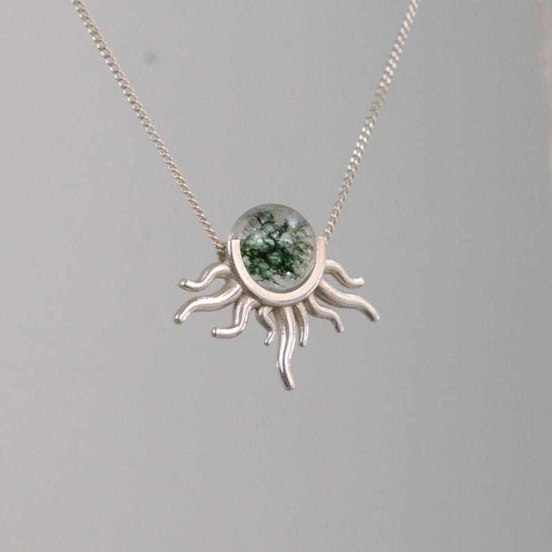 K&ouml;nnte beinhalten: Eine silberne Halskette mit einem sonnenf&ouml;rmigen Anh&auml;nger. Der Anh&auml;nger hat eine klare, runde Mitte mit gr&uuml;nen Einschl&uuml;ssen, eingebettet in ein silbernes Sonnendesign. Die Halskette ist ebenfalls aus Silber.