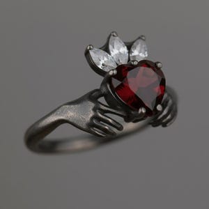 Peut inclure: Bague argentée ornée d'une pierre rouge en forme de cœur, tenue par deux mains. Au-dessus du cœur, une couronne de pierres blanches. La bague a une esthétique vintage.