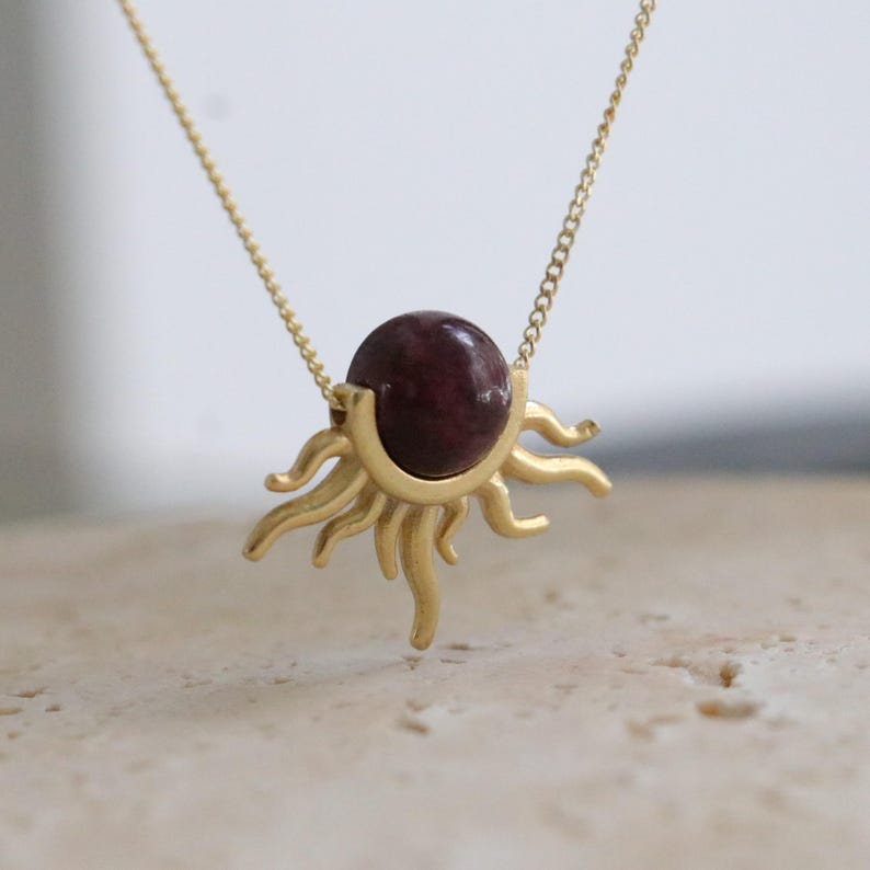 K&ouml;nnte beinhalten: Goldfarbene Halskette mit einem Sonnenanh&auml;nger und einem runden, dunkelroten Edelstein in der Mitte. Die Sonnenstrahlen sind stilisiert und gebogen. Die Kette ist filigran.
