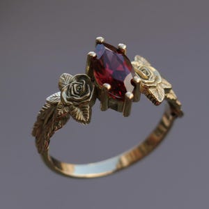 Könnte beinhalten: Ein goldfarbener Ring mit einem tiefroten, marquiseförmigen Edelstein. Der Ring ist mit detaillierten Rosen- und Blattmotiven auf beiden Seiten des Edelsteins verziert, wodurch ein Blumenmotiv entsteht. Das Band des Rings ist glatt und poliert.